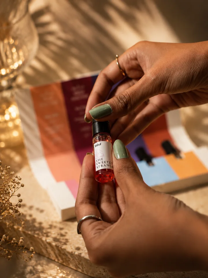 Hands with sage-green nails holding the Sweetest Stranger vial, its juice rose-pink, in front of the open kit and a crystal glass