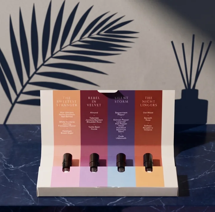 The Discovery Kit open on dark blue marble: four vials above pink, lilac, blue and peach panels, under a lid listing each fragrance's notes, with the shadows of a palm leaf and a reed diffuser on the wall