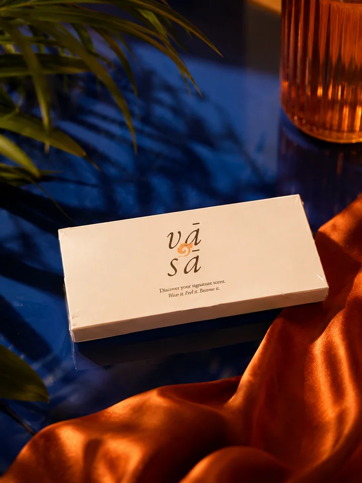 The Discovery Kit closed in its ivory sleeve with the vā sā mark, on dark blue glass beside copper satin, palm leaves and an amber glass