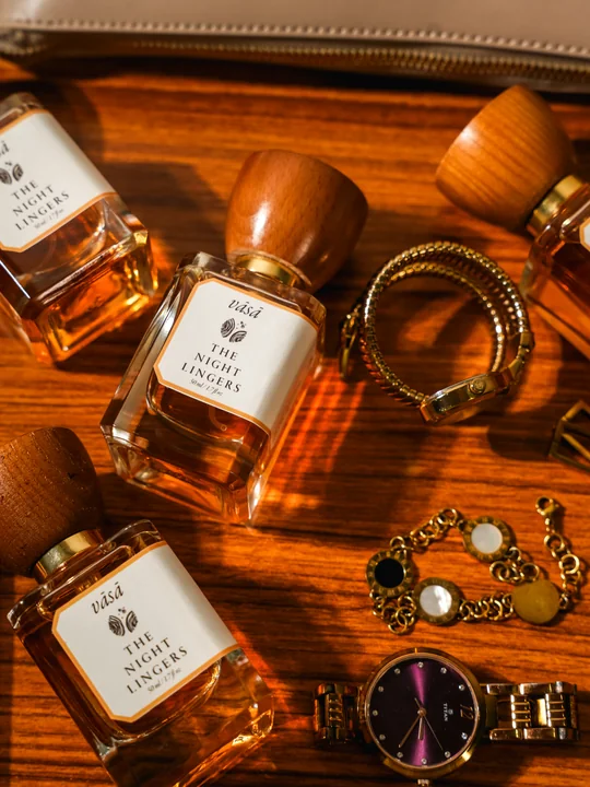 Bottles of The Night Lingers on a warm wooden table among gold bracelets and a watch